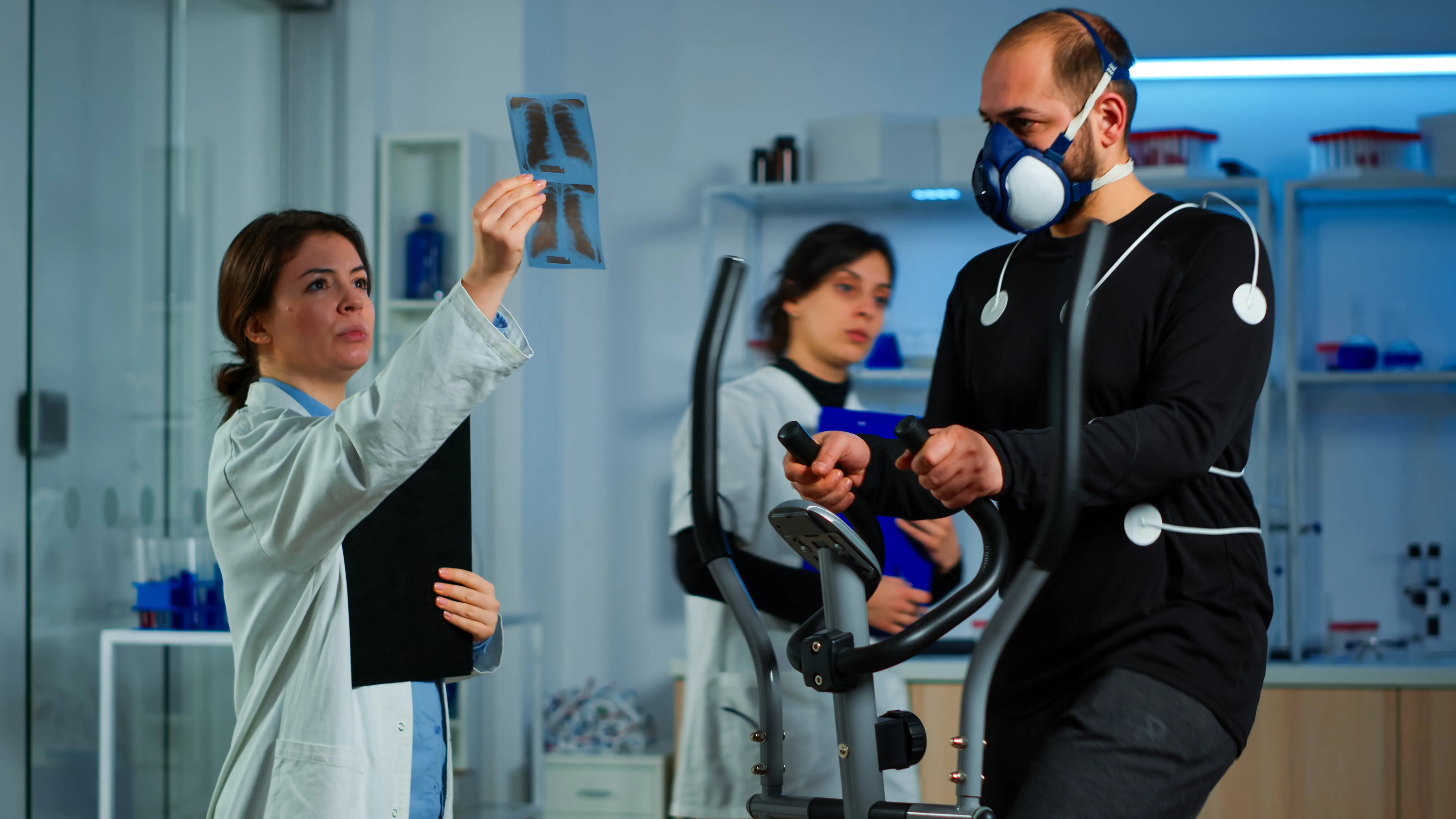 A medical professional in a white lab coat holds up and examines a chest X-ray while a male patient, wearing a respiratory mask and sensors on his chest, exercises on an elliptical machine in a clinical or laboratory setting. Another medical staff member stands in the background holding a blue clipboard